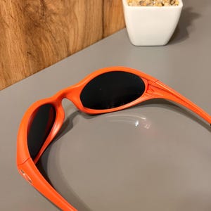May include: Bright orange sunglasses with black lenses. The frames are a vibrant, solid orange color, and the temples have a sleek, curved design. The lenses are dark, providing shade. The sunglasses are a sporty style.