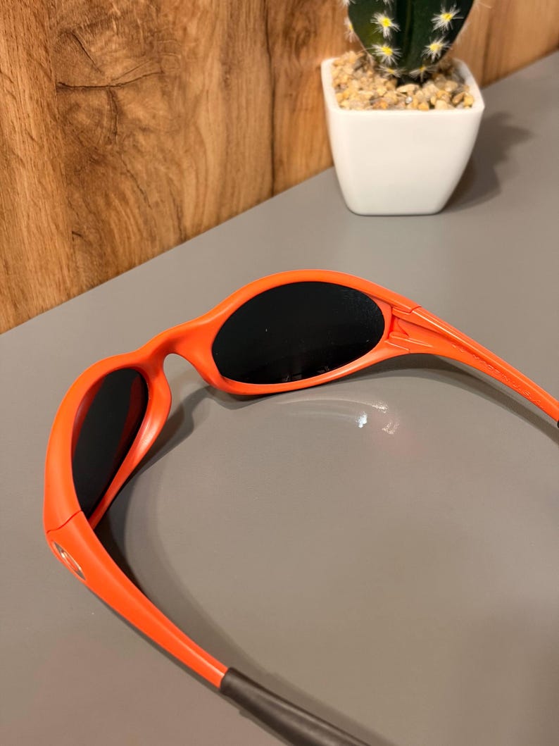May include: Bright orange sunglasses with black lenses. The frames are a vibrant, solid orange color, and the temples have a sleek, curved design. The lenses are dark, providing shade. The sunglasses are a sporty style.