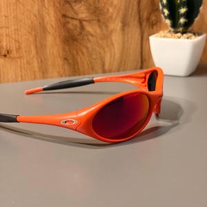 May include: Orange sunglasses with red-tinted lenses. The sunglasses have a sporty design with a black accent on the temples. The lenses are oval-shaped and the frame is a bright orange color.