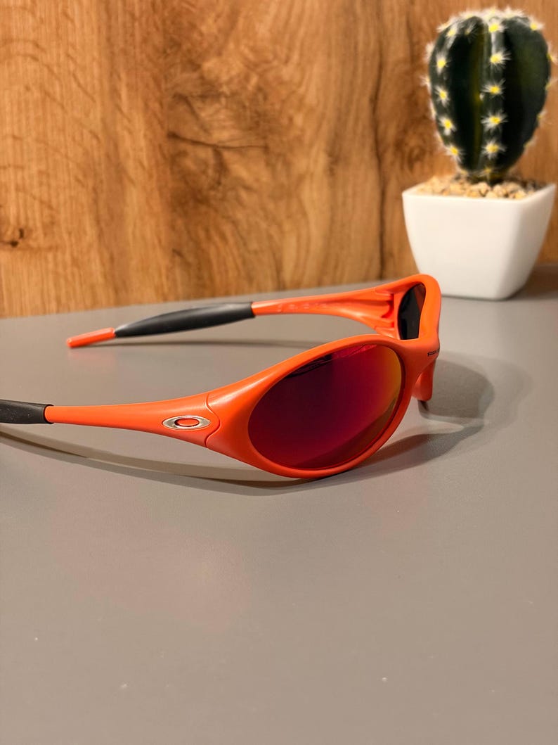 May include: Orange sunglasses with red-tinted lenses. The sunglasses have a sporty design with a black accent on the temples. The lenses are oval-shaped and the frame is a bright orange color.
