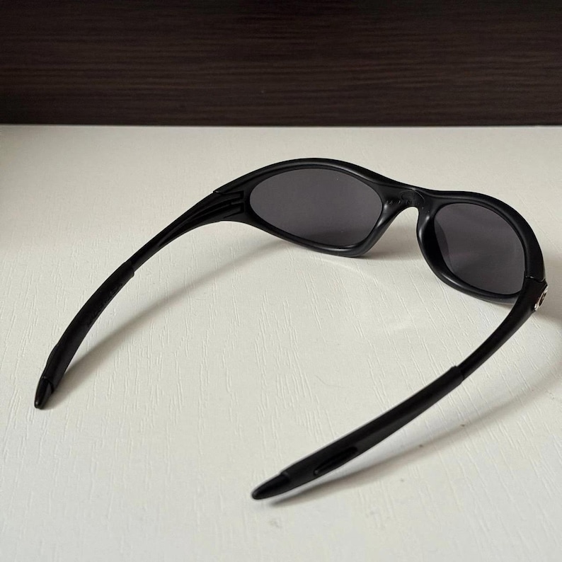 Puede incluir: Gafas de sol negras con lentes de color gris oscuro. Las gafas de sol tienen un dise&ntilde;o elegante y deportivo con una montura y patillas curvas. Las lentes son de forma ovalada.