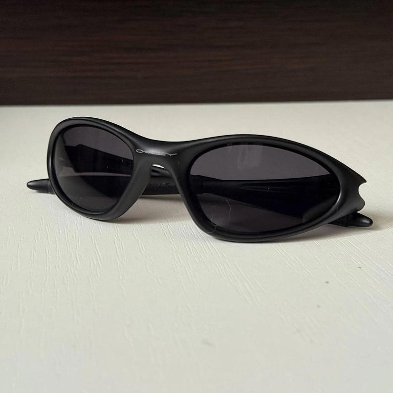 Puede incluir: Gafas de sol negras con montura curvada y lentes oscuras. Las gafas de sol tienen un dise&ntilde;o deportivo y est&aacute;n sobre una superficie blanca. El nombre de la marca es visible en la montura.
