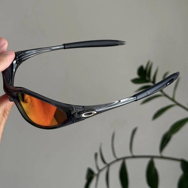 Puede incluir: Gafas de sol negras y transparentes con lentes naranjas. Las gafas tienen un dise&ntilde;o elegante y moderno con una montura curvada y patillas oscuras. Las lentes son de color naranja vibrante.