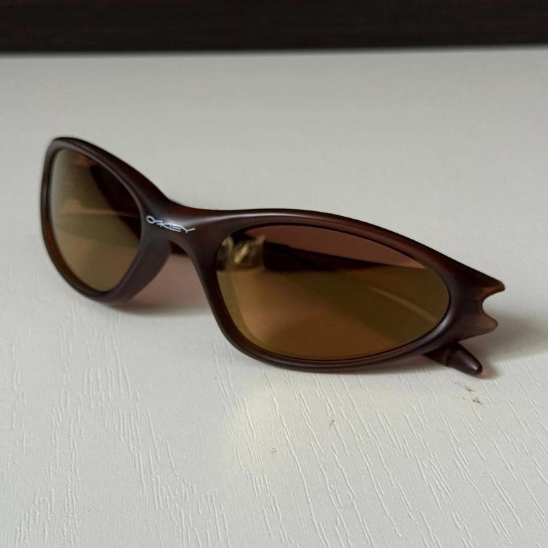 May include: Brown sunglasses with a curved frame and tinted lenses. The brand name "Oakley" is visible on the left arm. The sunglasses are designed with a sleek, sporty aesthetic.