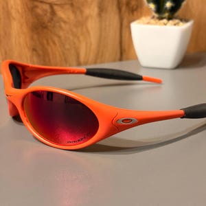 May include: Orange sunglasses with red-tinted lenses. The sunglasses have a sporty design with a curved frame and black earpieces. The lenses are oval-shaped and feature the word "PRIZM" printed on the lens.