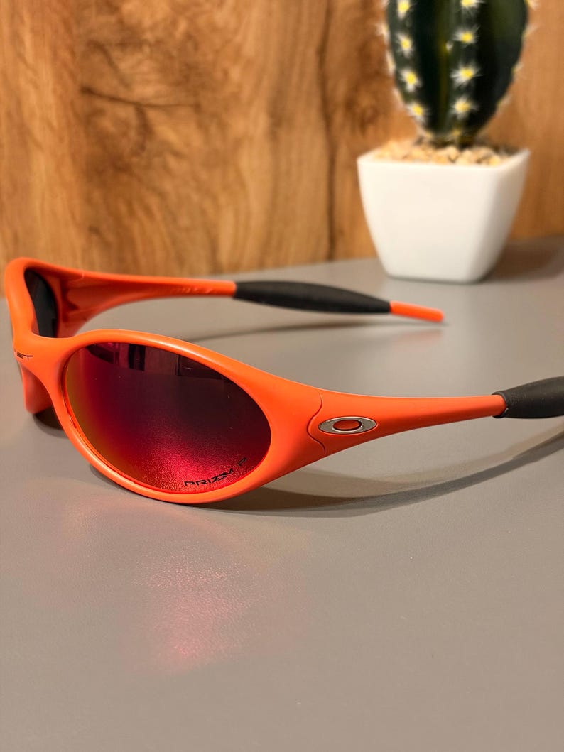 May include: Orange sunglasses with red-tinted lenses. The sunglasses have a sporty design with a curved frame and black earpieces. The lenses are oval-shaped and feature the word "PRIZM" printed on the lens.