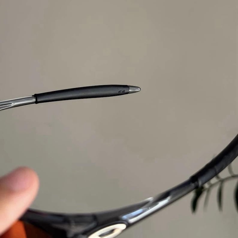 Puede incluir: Primer plano de gafas de sol negras con montura plateada y patilla negra. La patilla tiene una punta redondeada y una peque&ntilde;a marca. Las gafas de sol son parcialmente visibles sobre un fondo neutro.