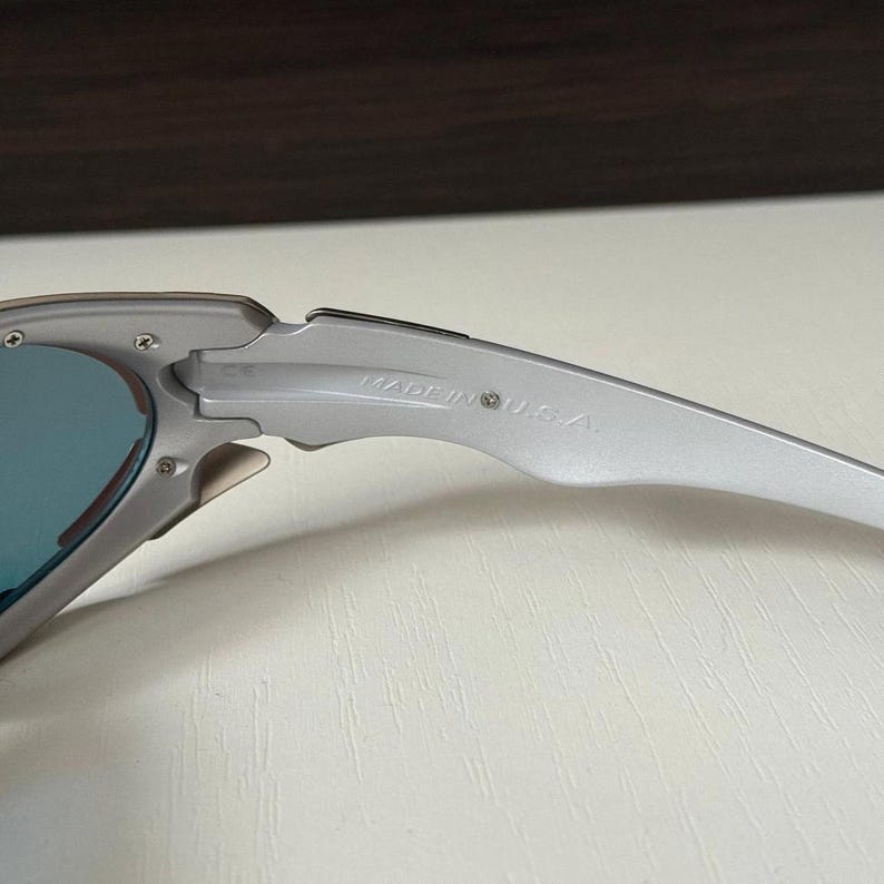 Puede incluir: Primer plano de gafas de sol plateadas con lentes azules. La montura met&aacute;lica lleva la inscripci&oacute;n "MADE IN U.S.A." en la patilla. Las lentes tienen una calidad reflectante.