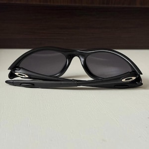 Puede incluir: Gafas de sol negras con montura curvada y lentes oscuras. Las gafas de sol est&aacute;n plegadas, revelando las bisagras y el interior de las patillas. Las lentes son de forma ovalada.
