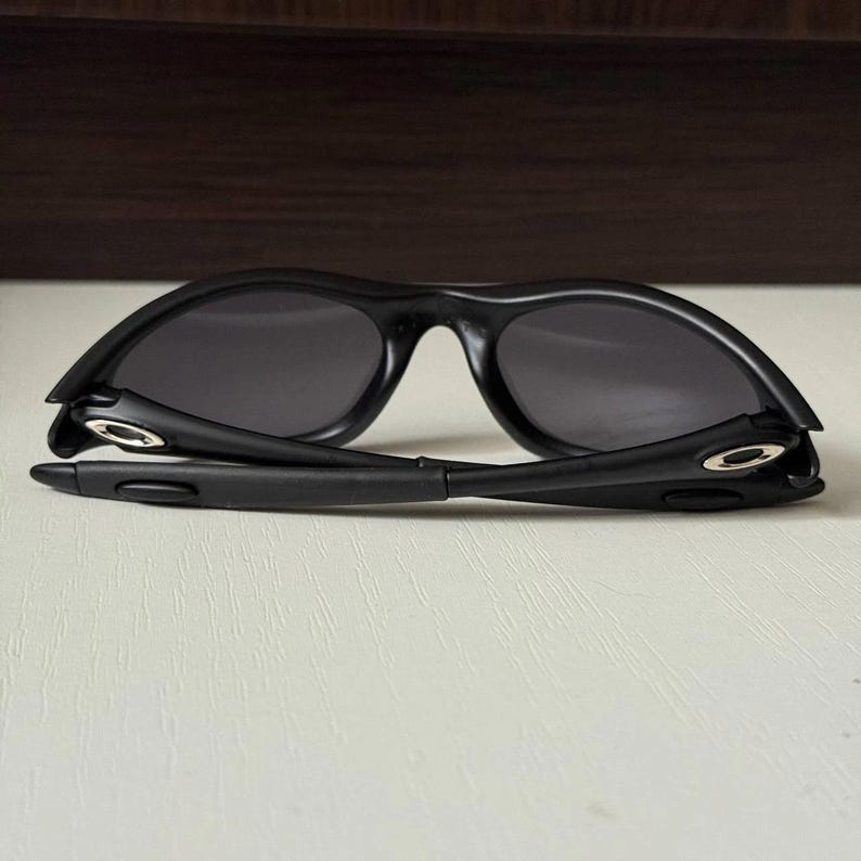 Puede incluir: Gafas de sol negras con montura curvada y lentes oscuras. Las gafas de sol est&aacute;n plegadas, revelando las bisagras y el interior de las patillas. Las lentes son de forma ovalada.