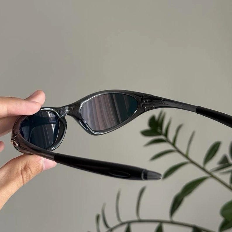 Puede incluir: Gafas de sol negras con montura curvada y lentes oscuras. Las lentes reflejan una mezcla de colores. Las patillas son elegantes y se extienden hacia atr&aacute;s, creando un aspecto moderno y deportivo. Las gafas de sol se sostienen, mostrando su dise&ntilde;o.
