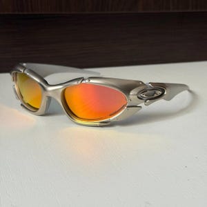 Puede incluir: Gafas de sol plateadas con lentes naranjas y amarillas. Las gafas tienen un dise&ntilde;o deportivo con una montura curvada y un logotipo en la patilla. Las lentes tienen un efecto degradado, pasando del naranja al amarillo.