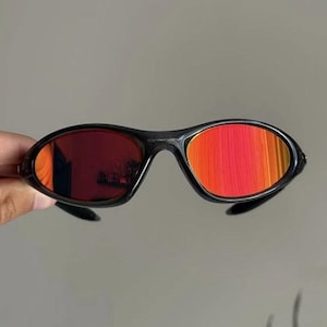 Puede incluir: Gafas de sol negras con lentes rojo-naranja. Las gafas tienen una montura elegante y curvada y un dise&ntilde;o deportivo. Las lentes reflejan un degradado de colores, a&ntilde;adiendo un toque vibrante a las gafas.