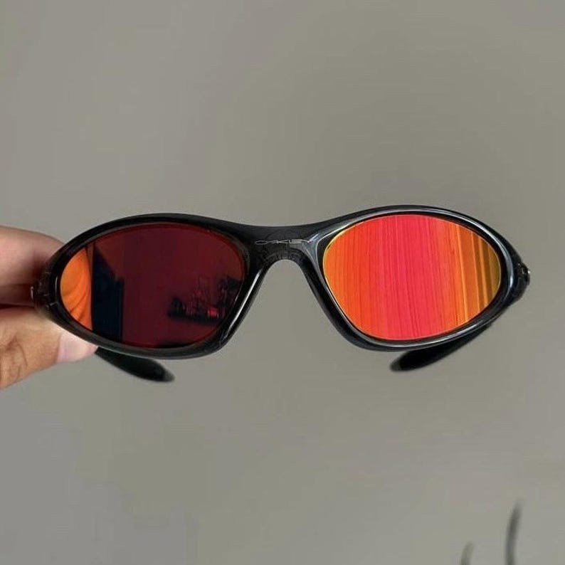 Puede incluir: Gafas de sol negras con lentes rojo-naranja. Las gafas tienen una montura elegante y curvada y un dise&ntilde;o deportivo. Las lentes reflejan un degradado de colores, a&ntilde;adiendo un toque vibrante a las gafas.