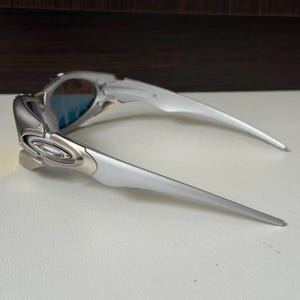 Puede incluir: Gafas de sol plateadas y grises con lentes tintadas en azul. Las gafas de sol tienen un dise&ntilde;o elegante y moderno con patillas curvas y un acabado met&aacute;lico. Las lentes son reflectantes y la montura tiene una estructura angular detallada.