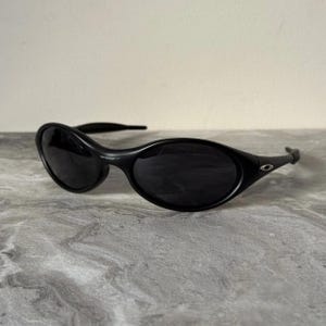 Puede incluir: Gafas de sol negras de forma ovalada con lentes oscuras. Las gafas tienen una montura y patillas curvas. El logotipo de Oakley es visible en la montura. Las gafas están sobre una superficie gris.