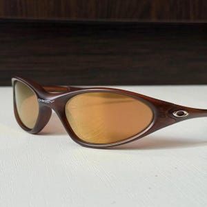 May include: Brown Oakley sunglasses with gold-tinted lenses. The sunglasses have a curved frame and the Oakley logo is visible on the left arm. The lenses have a reflective quality.