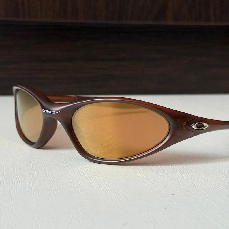 May include: Brown Oakley sunglasses with gold-tinted lenses. The sunglasses have a curved frame and the Oakley logo is visible on the left arm. The lenses have a reflective quality.