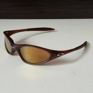 May include: Brown sunglasses with a curved frame and gold-tinted lenses. The Oakley logo is visible on the frame. The sunglasses are designed for sports and outdoor activities.