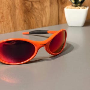 May include: Bright orange sunglasses with red-tinted lenses. The sporty eyewear features a curved frame and dark gray temples for a secure fit. The sunglasses are set against a neutral background with a small cactus in a white pot.