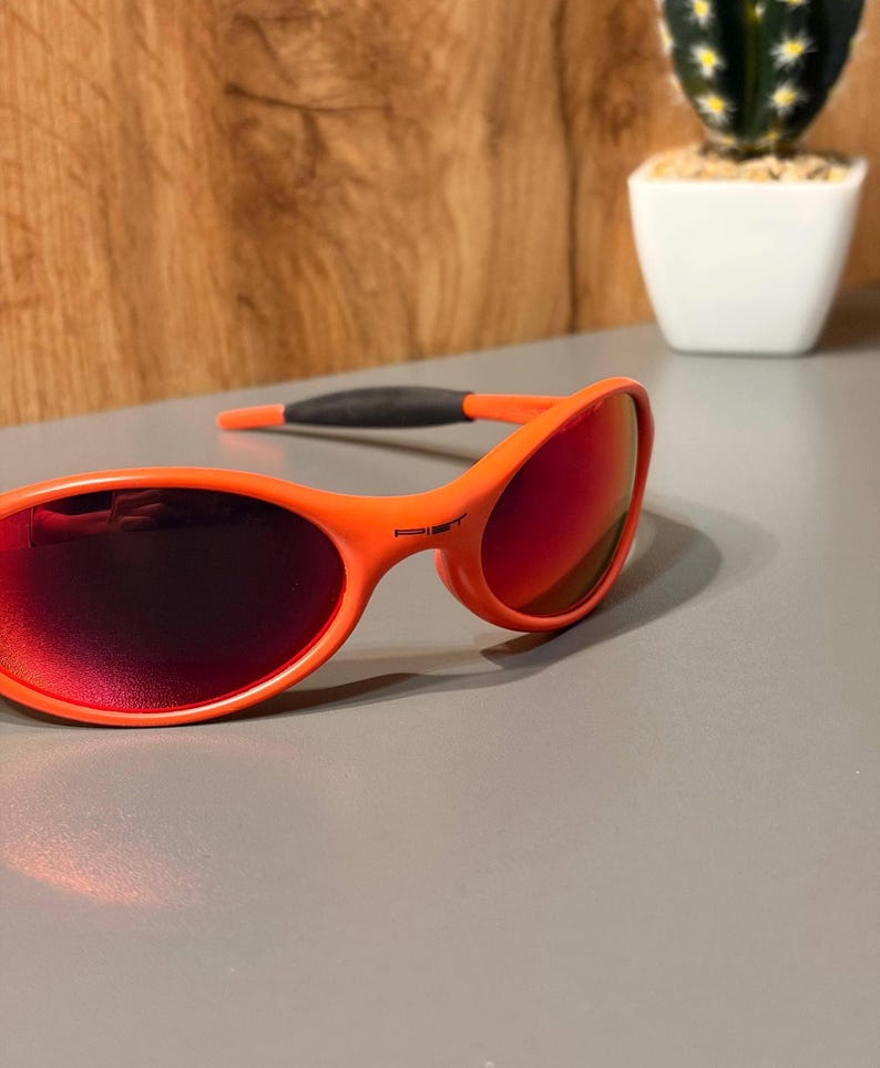 May include: Bright orange sunglasses with red-tinted lenses. The sporty eyewear features a curved frame and dark gray temples for a secure fit. The sunglasses are set against a neutral background with a small cactus in a white pot.