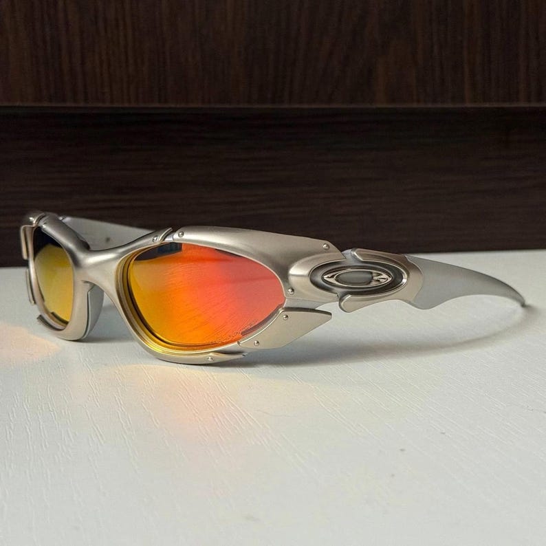Puede incluir: Gafas de sol plateadas con lentes naranjas y amarillas. Las gafas de sol tienen un dise&ntilde;o futurista con una montura curvada y una est&eacute;tica elegante y moderna. Las lentes reflejan un degradado de colores.