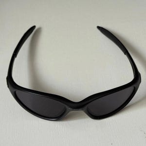 Puede incluir: Gafas de sol negras con lentes oscuras. Las gafas de sol tienen un dise&ntilde;o elegante y moderno con una montura y patillas curvas. Las lentes son de color gris oscuro y la montura tiene un acabado negro mate. Las gafas est&aacute;n dise&ntilde;adas para la protecci&oacute;n ocular.