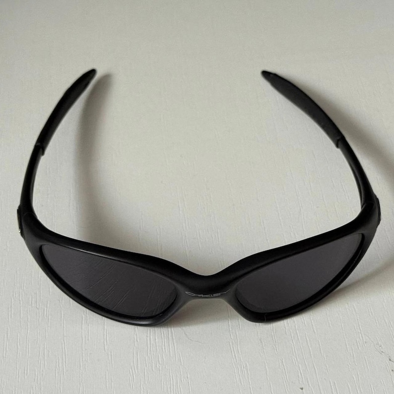 Puede incluir: Gafas de sol negras con lentes oscuras. Las gafas de sol tienen un dise&ntilde;o elegante y moderno con una montura y patillas curvas. Las lentes son de color gris oscuro y la montura tiene un acabado negro mate. Las gafas est&aacute;n dise&ntilde;adas para la protecci&oacute;n ocular.