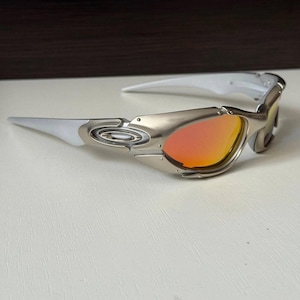 Puede incluir: Gafas de sol plateadas y blancas con lentes naranjas. Las gafas tienen un dise&ntilde;o elegante y moderno con montura met&aacute;lica y patillas blancas. Las lentes son de color naranja vibrante, ofreciendo un contraste audaz con la montura plateada.
