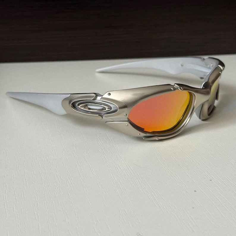 Puede incluir: Gafas de sol plateadas y blancas con lentes naranjas. Las gafas tienen un dise&ntilde;o elegante y moderno con montura met&aacute;lica y patillas blancas. Las lentes son de color naranja vibrante, ofreciendo un contraste audaz con la montura plateada.