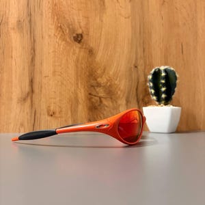 May include: Orange and black sports sunglasses with red mirrored lenses. The sunglasses are set against a neutral gray surface with a wooden background and a small cactus in a white pot.