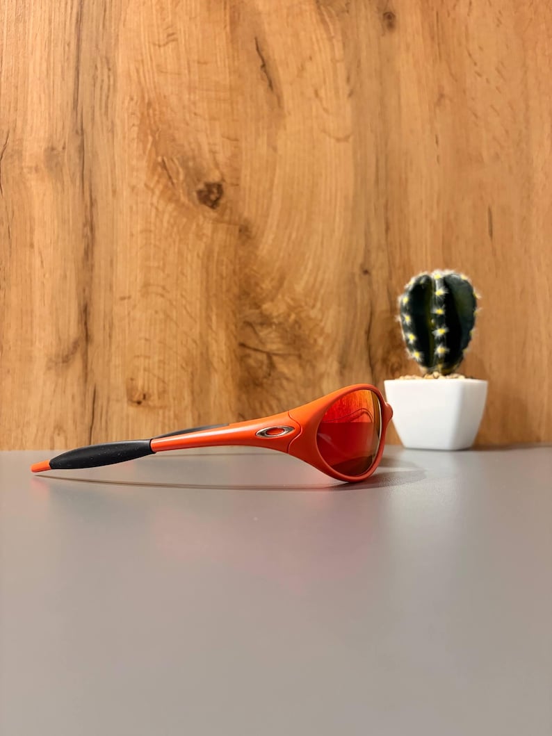 May include: Orange and black sports sunglasses with red mirrored lenses. The sunglasses are set against a neutral gray surface with a wooden background and a small cactus in a white pot.