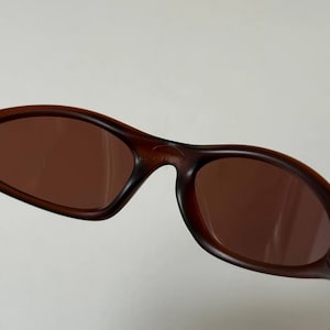 May include: Brown sunglasses with a curved frame and brown lenses. The sunglasses have a sleek, sporty design, suitable for outdoor activities. The lenses appear to be tinted, offering protection from the sun.