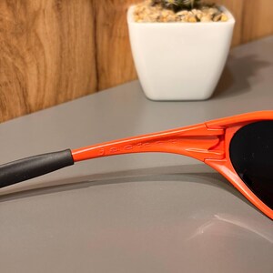 May include: Close-up of orange sunglasses with black lenses and the word "jacket" printed on the arm. The sunglasses have a sleek, modern design. The arm of the sunglasses is orange with a black tip.