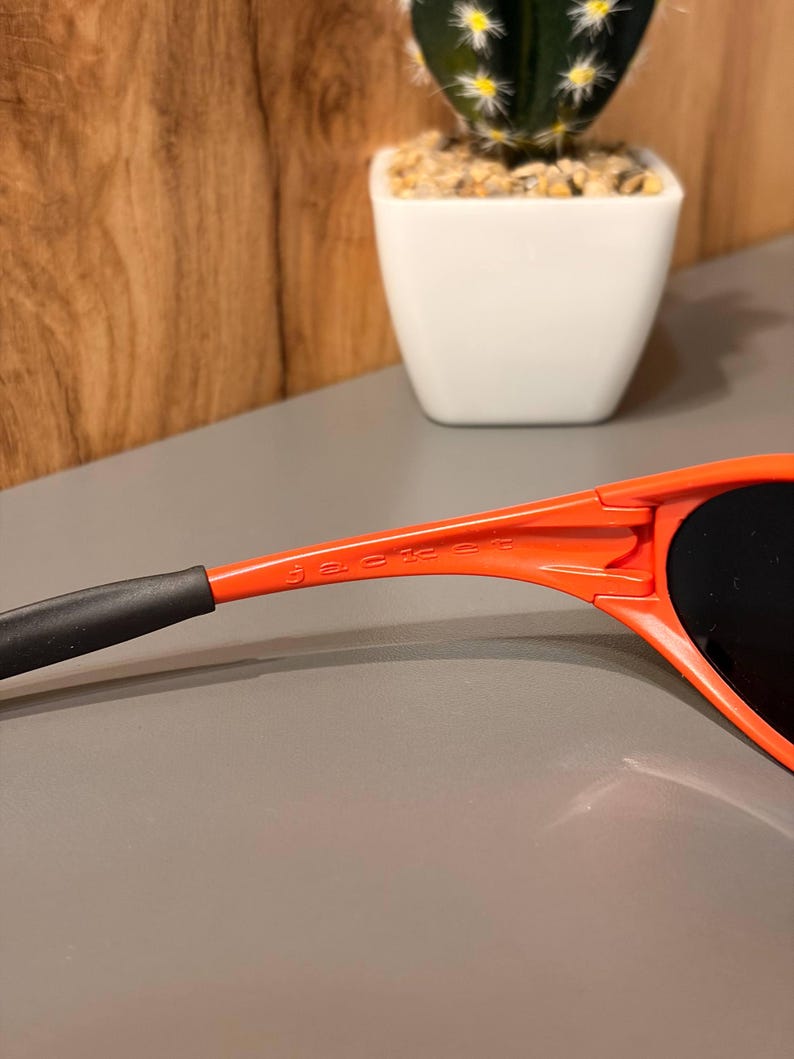 May include: Close-up of orange sunglasses with black lenses and the word "jacket" printed on the arm. The sunglasses have a sleek, modern design. The arm of the sunglasses is orange with a black tip.