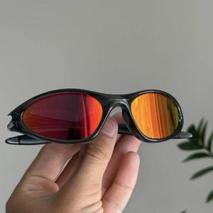 Puede incluir: Gafas de sol negras con lentes degradadas rojas y naranjas. Las gafas tienen un dise&ntilde;o de montura elegante y curvado. Las lentes reflejan un vibrante espectro de colores, creando un look audaz y elegante.