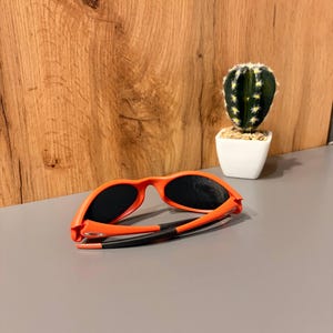 May include: Orange sunglasses with black lenses and black arms are displayed on a gray surface. The sunglasses are folded. A small cactus in a white pot is in the background.
