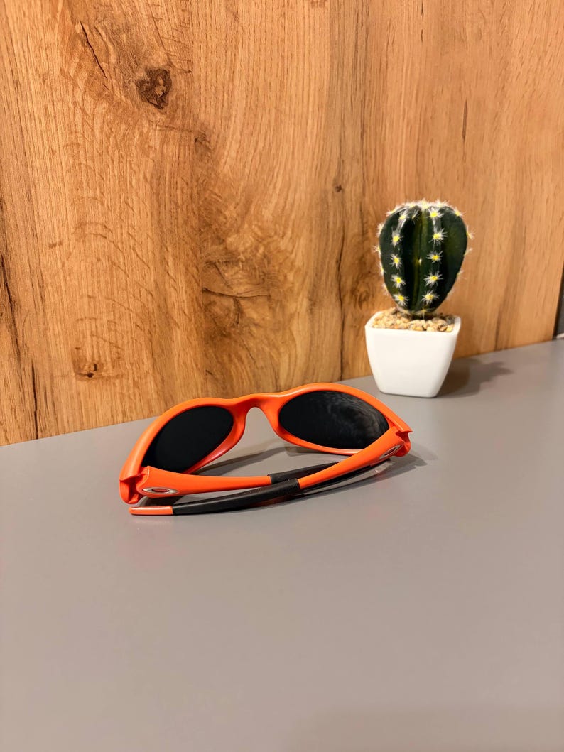 May include: Orange sunglasses with black lenses and black arms are displayed on a gray surface. The sunglasses are folded. A small cactus in a white pot is in the background.