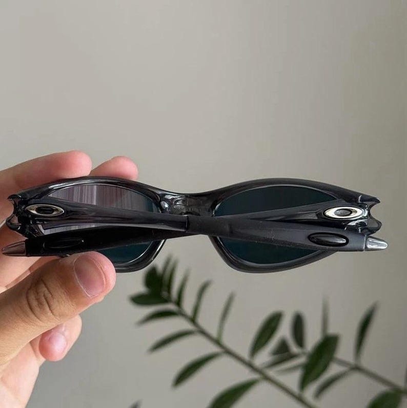 Puede incluir: Gafas de sol negras con un dise&ntilde;o elegante y moderno. Las gafas tienen una montura curvada y lentes oscuras. Las patillas de las gafas tienen un detalle plateado. Las gafas son sostenidas por una mano.