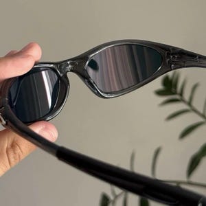 Puede incluir: Gafas de sol negras con montura curvada y lentes oscuras. Las gafas de sol tienen un dise&ntilde;o elegante y moderno, con un acabado brillante. Las lentes reflejan la luz, creando un efecto de espejo. Las patillas son finas.