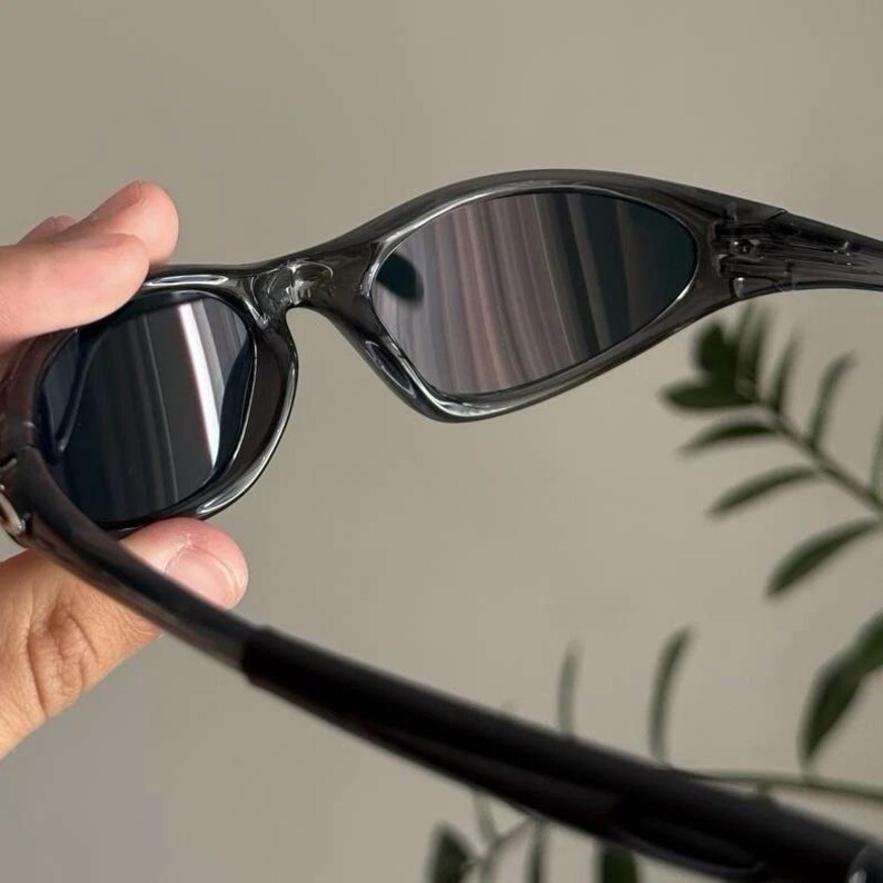 Puede incluir: Gafas de sol negras con montura curvada y lentes oscuras. Las gafas de sol tienen un dise&ntilde;o elegante y moderno, con un acabado brillante. Las lentes reflejan la luz, creando un efecto de espejo. Las patillas son finas.