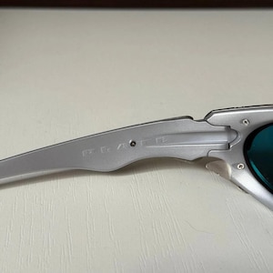 Puede incluir: Primer plano de gafas de sol plateadas con lentes azules. La montura tiene un acabado met&aacute;lico y un dise&ntilde;o elegante y moderno. Las gafas de sol est&aacute;n en &aacute;ngulo, mostrando los detalles de la montura y las lentes.