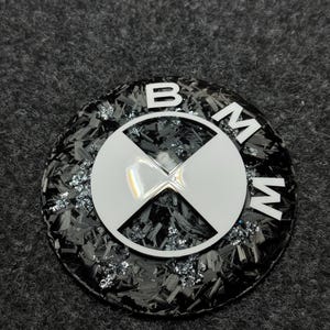 Puede incluir: Un emblema redondo de BMW con un fondo de fibra de carbono negra y motas plateadas. El logotipo blanco de BMW presenta las letras "B", "M" y "W" alrededor de una cruz blanca.