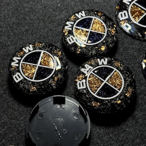 May include: A collection of black and gold BMW wheel center caps. The caps feature the BMW logo in white, with a gold and navy blue design. The caps have a textured appearance and are scattered on a dark surface.