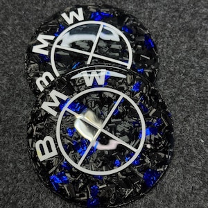 May include: Two black and blue BMW wheel center caps. The caps have a carbon fiber appearance with blue accents. The BMW logo is white and features the letters "BMW" in a circular design.