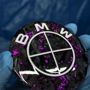 May include: A round, black and purple BMW emblem with the letters "BMW" in white. The emblem has a carbon fiber appearance with purple accents. The background is a blurred blue.