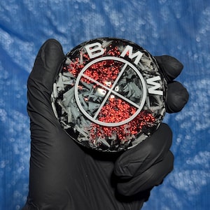 May include: A round, black and gray emblem with the letters "BMW" in white. The center features a red glitter design. The emblem is held by a gloved hand against a blue background.