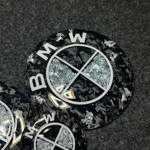 May include: Close-up of several black and silver BMW emblem car accessories. The emblems feature the BMW logo in white, with a silver and black carbon fiber pattern. The background is a dark gray surface.