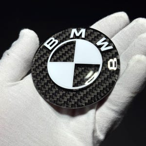 May include: A circular BMW emblem featuring a black and white checkered pattern with a carbon fiber texture. The outer ring displays the letters B, M, and W in white against a dark background. The design is glossy and reflective in appearance.