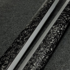 Puede incluir: Dos piezas de moldura automotriz largas y estrechas con un acabado brillante de fibra de carbono forjada con un patrón jaspeado negro y gris. Cada pieza tiene un borde gris mate texturizado a lo largo de un lado para contrastar.